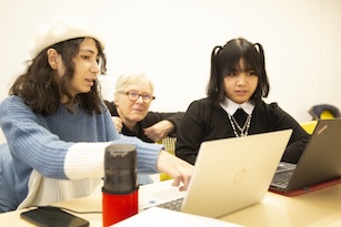An instructor kneels down and interacts with 2 students who are working with laptops at a table.